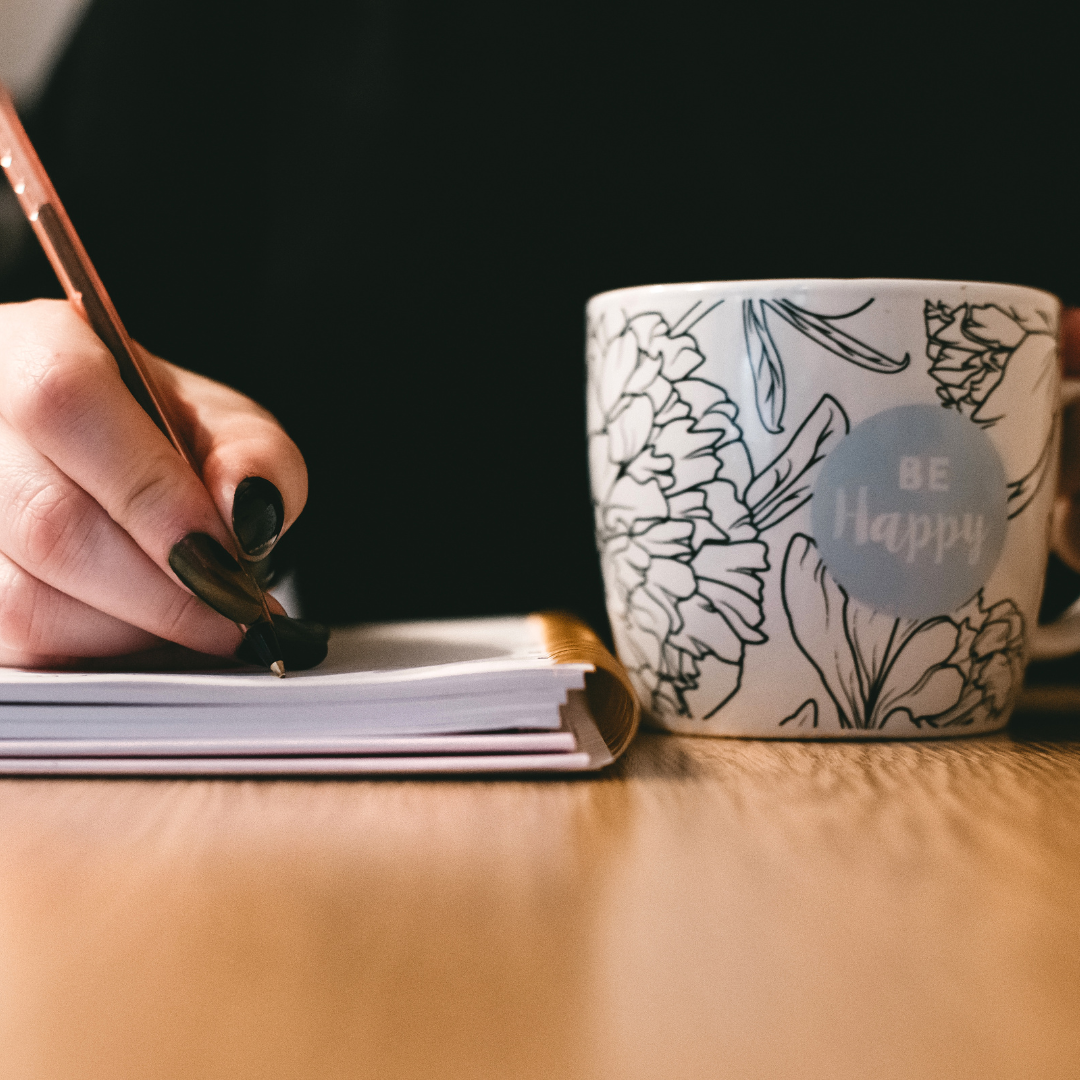 Close-up of a hand writing in a notebook beside a floral mug with the words 'Be Happy,' symbolizing creativity, reflection, and the personalized branding focus of the Epic Experience package.