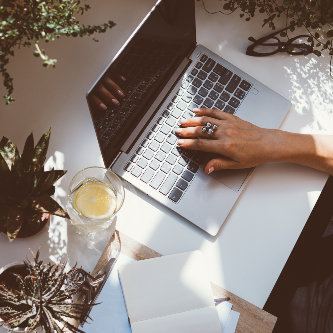 A person working on a laptop at a desk surrounded by plants and a glass of lemon water, showcasing the fresh and creative environment fostered by the Novel Narrative package.