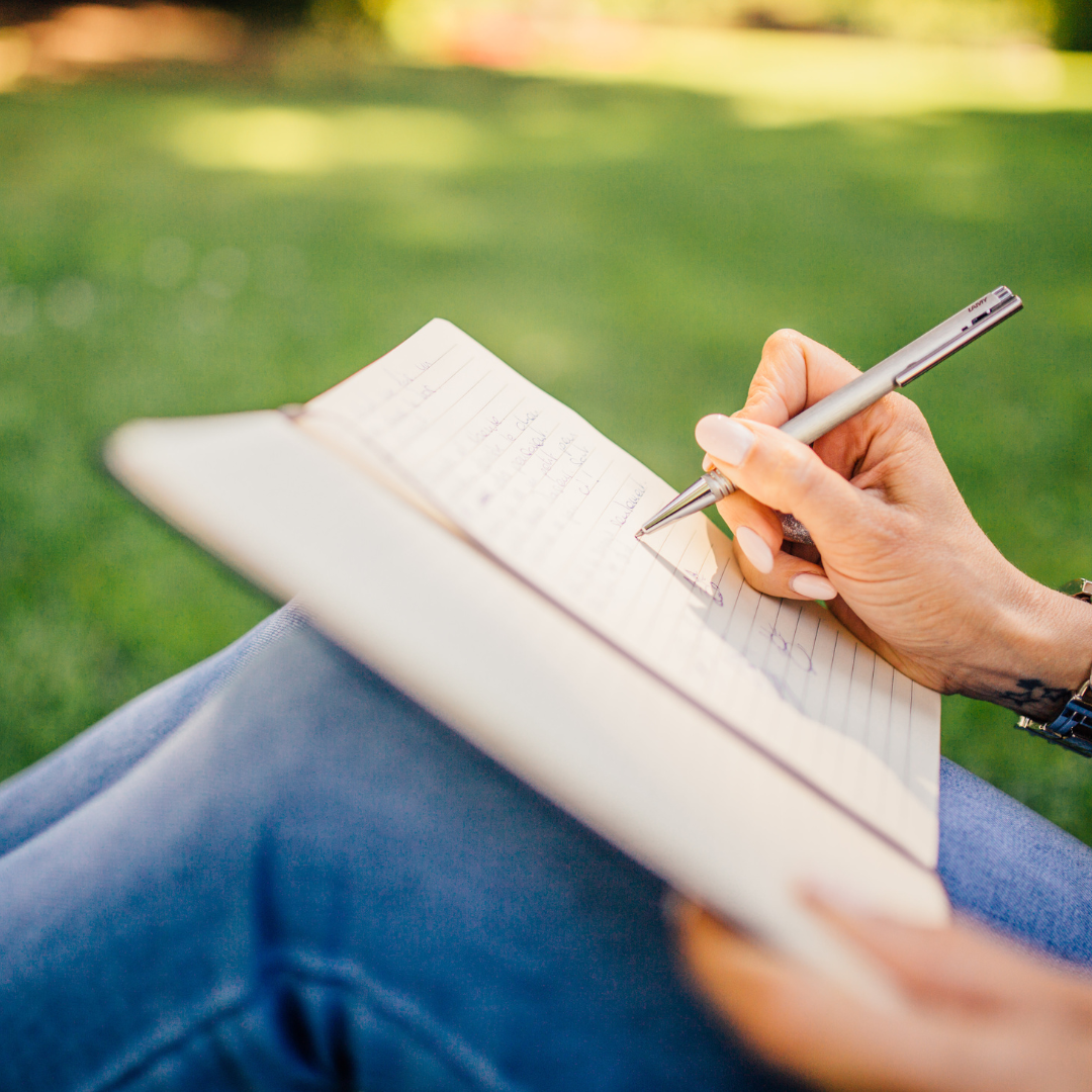 Close-up of a person writing in a notebook while sitting outdoors on green grass, embodying the fresh start and personalized strategies provided in the Storyline Success package.