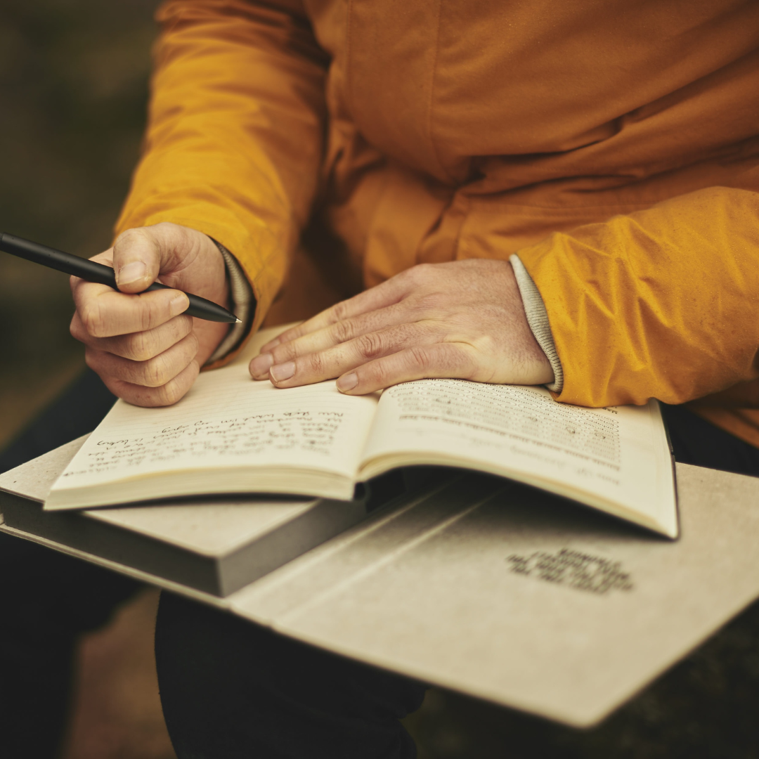 Close-up of a person writing in a notebook outdoors, wearing a bright orange jacket, symbolizing focused effort and the hands-on branding support of the Work in Progress package.